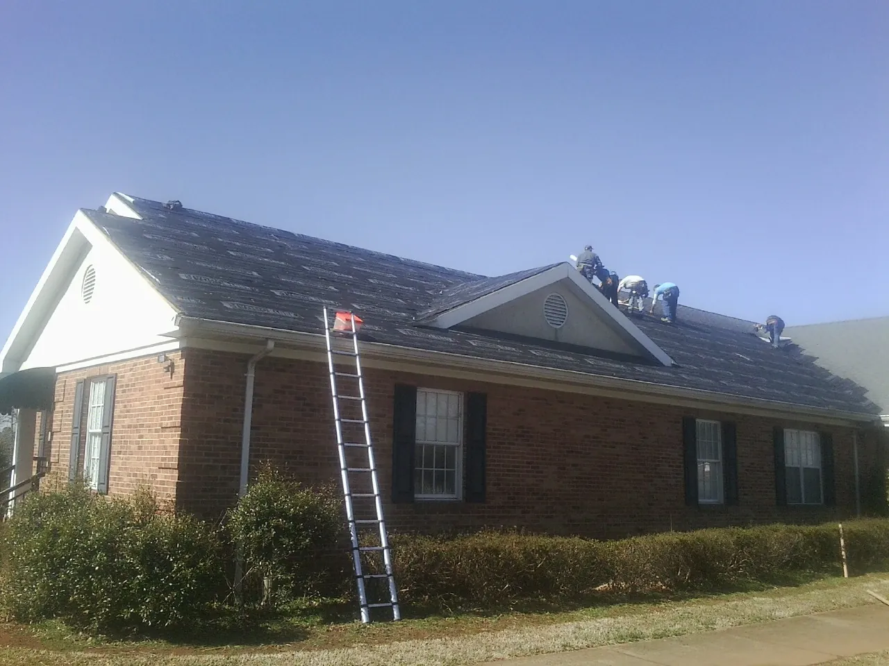 Crew tearing shingles off a brick ranch roof with a ladder set — Emergency Roof Repair work by our crew in Hardy