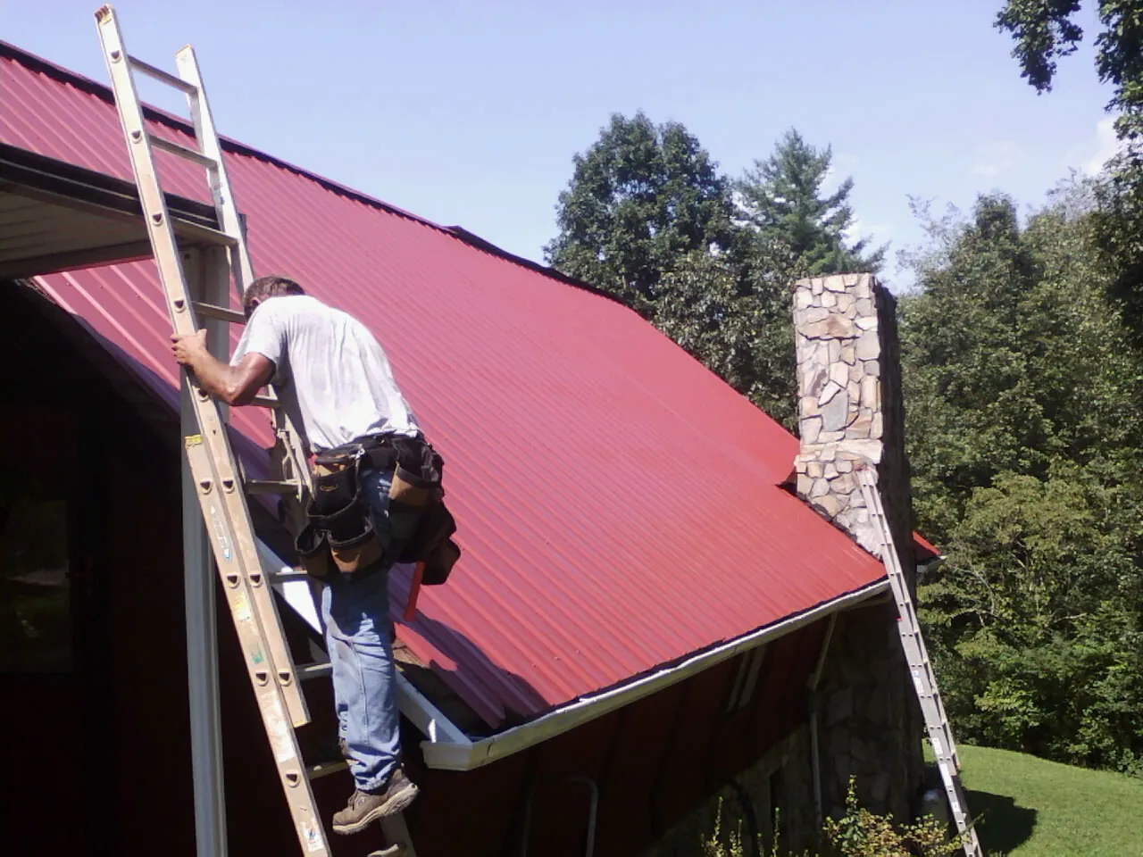Roofer on a ladder beside a red metal roof and stone chimney — Soffit & Fascia Repair by Roofing Brothers in Hardy